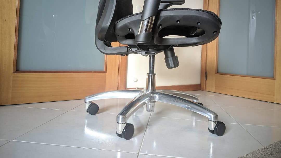 The lower part of an office chair, showing its polished aluminium five-star base, chrome gas lift and five black castor wheels on a tiled floor.