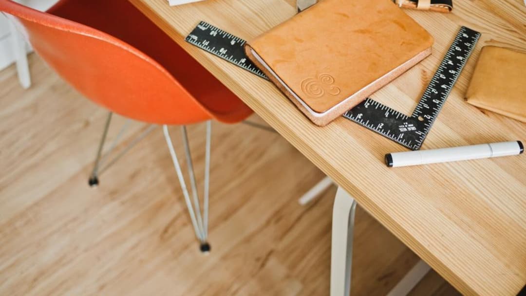 An overhead view of a light wooden desk with an orange moulded chair on thin metal legs, a leather notebook, two black rulers and a white pen.