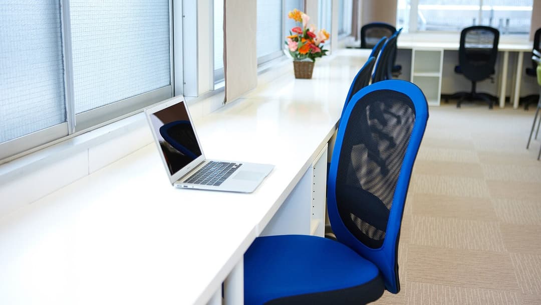 A blue mesh-backed office chair at a long white desk by a window with blinds, an open laptop on the desk and more office chairs along the room's carpeted floor behind it.