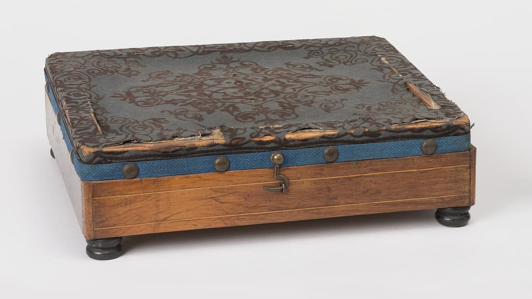 An old upholstered footstool with a patterned fabric top, blue trim and brass studs on a wooden base with rounded feet, on a plain background.