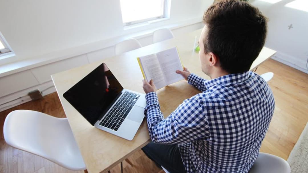 Seen from above, a person in a checked shirt sits at a light wooden table reading a booklet beside an open laptop.