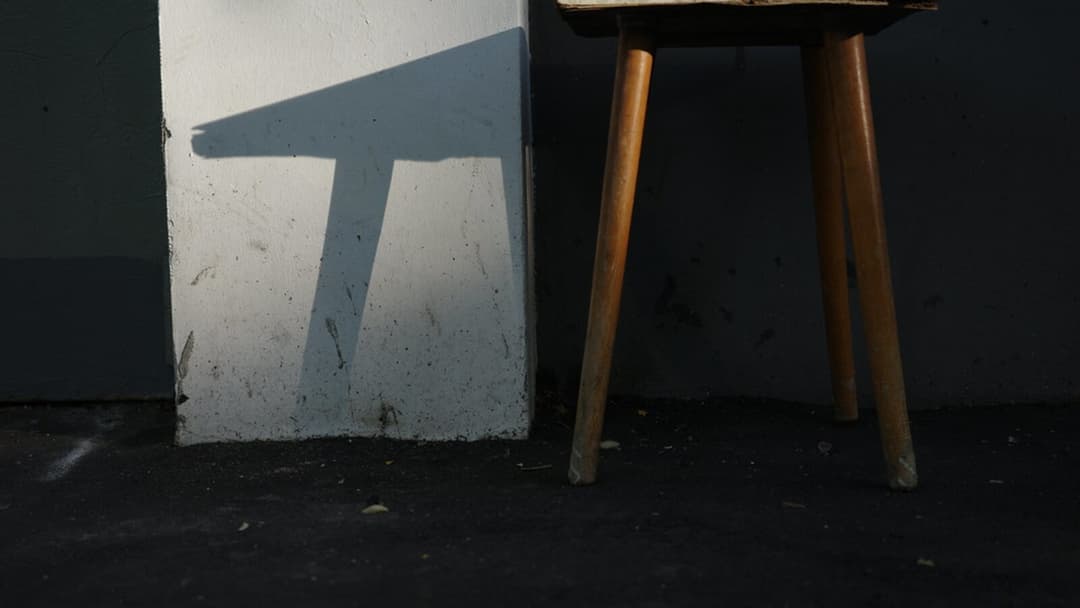 The legs and underside of a plain wooden stool casting an angular shadow on a pale wall.