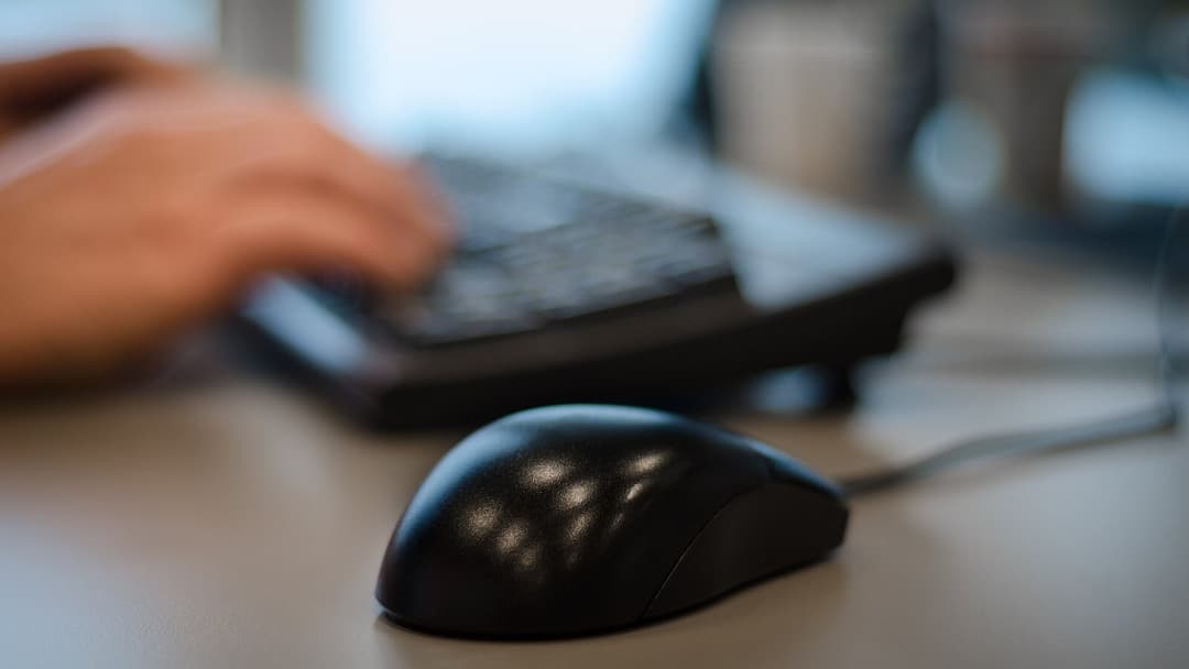 A hand resting on a black ergonomic keyboard with a black wired computer mouse in the foreground on a grey desk, with two blurred cups behind.