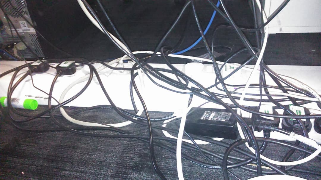 A dense tangle of black and white power cables looping over a white extension power strip and a black power adapter on a dark carpet beneath a desk.