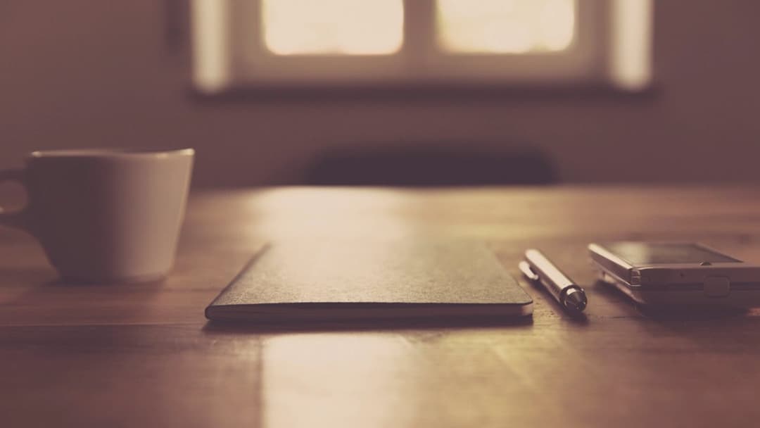 A white coffee mug, a closed notebook, a pen and a phone resting on a warm wooden desk, with a softly blurred window in the background.