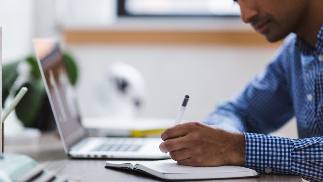 A person in a blue checked shirt writing in a notebook with a pen, an open laptop to the left, forearm resting on a wooden desk.