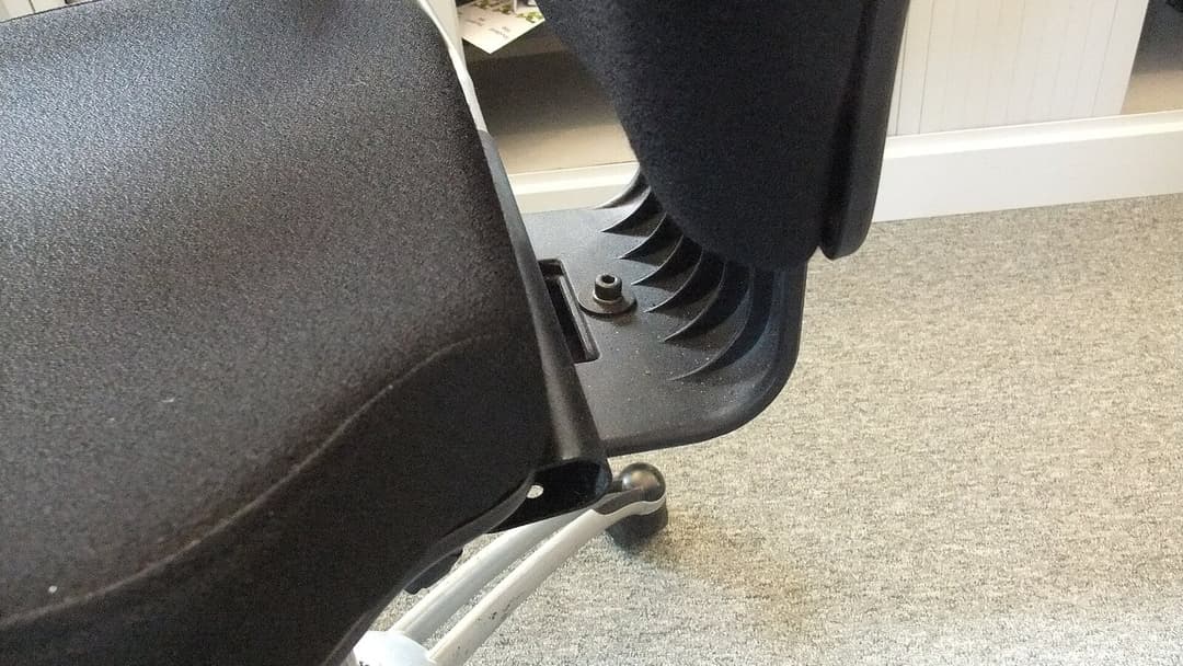 Close-up of the underside of a black office chair, showing the seat pan, the tilt mechanism, an adjustment knob and part of the metal star base on grey carpet.