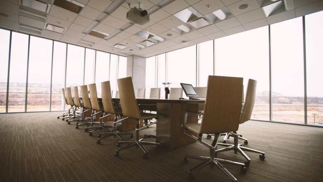 A row of high-backed office chairs with wood-veneer backs on chrome castor bases, lined along a long boardroom table between floor-to-ceiling windows.