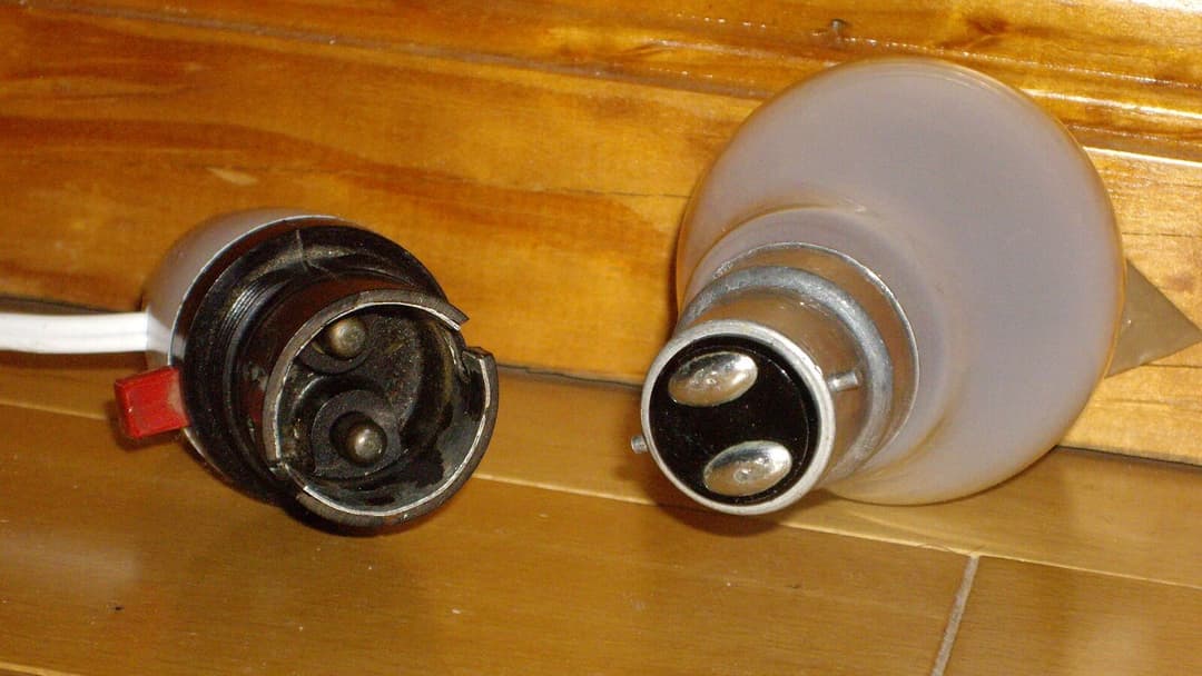 A frosted bulb with a metal bayonet base showing two round contacts lies on a wooden surface beside the matching open lamp holder it twists into, with a red switch on the holder.