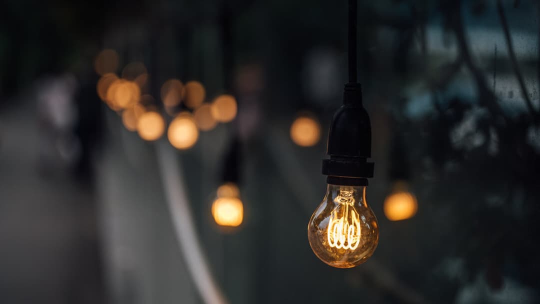 A clear glass bulb with a glowing curved filament hangs from a short black cord fitting in the dark, with several warm, out-of-focus lights strung away behind it.