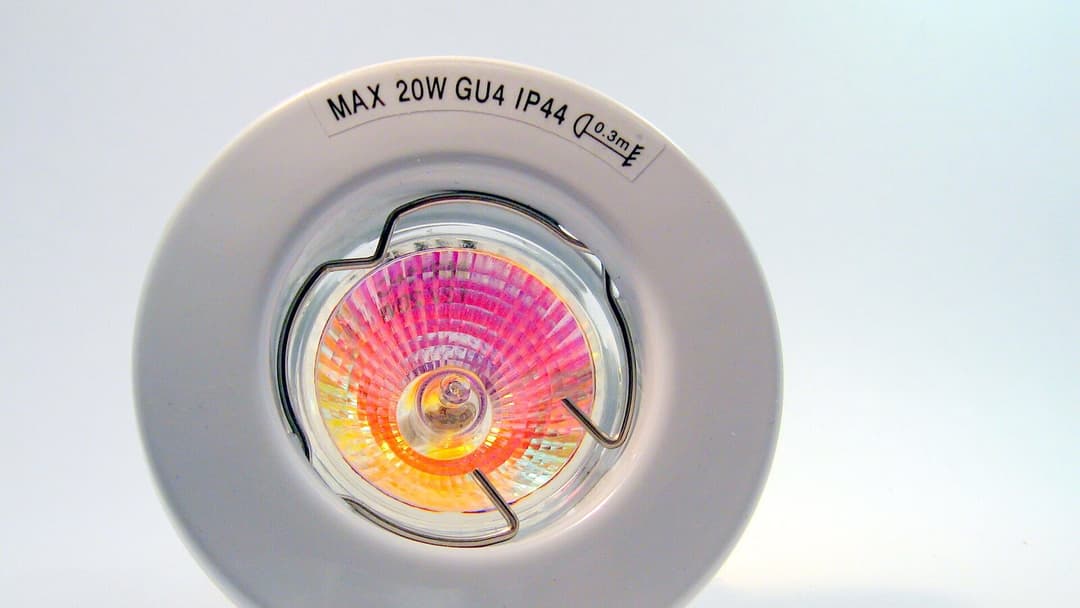 A round recessed spotlight fitting seen head-on, a colourful dichroic reflector lamp clipped into its centre, with a printed label on the white rim reading MAX 20W GU4 IP44.