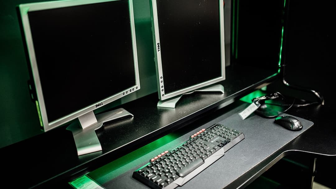 Two silver monitors with dark screens on a black desk shelf lit with green light, a black mechanical keyboard and a black mouse in front, with cables running off to the right.