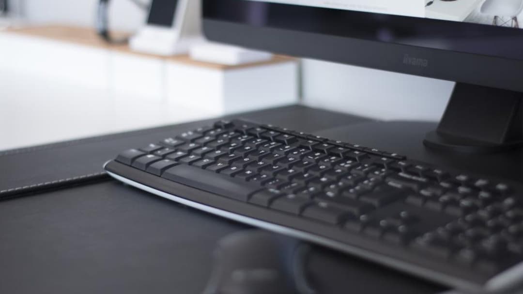 A black computer keyboard resting on a dark leather desk mat in front of a monitor with a dark screen, against a bright, out-of-focus desk.