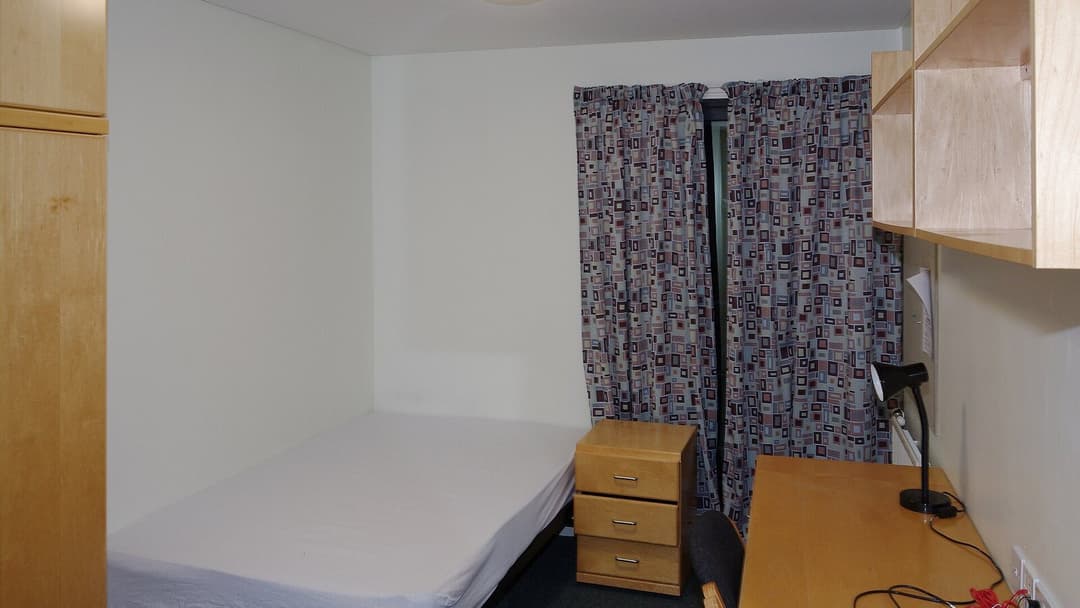A compact bedroom with a bare single bed, a wooden bedside drawer unit and patterned curtains, a tall wardrobe on the left, and a wooden desk with a black reading lamp along the right-hand wall.