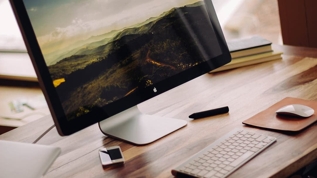 A desktop computer screen showing a mountain landscape on a wooden desk, with a wireless keyboard and a mouse on a leather mat to the right, and a stylus and a phone nearby.