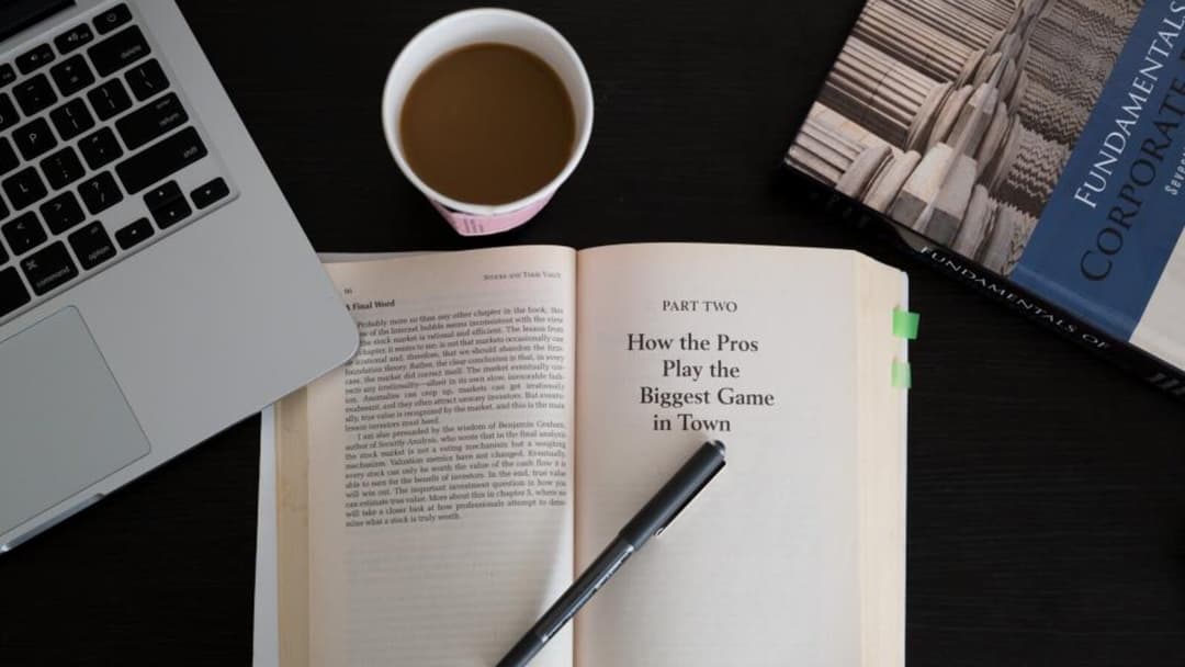 A flat lay on a dark desk with an open paperback and a pen resting on it, a cup of coffee, the corner of a laptop keyboard and a hardback book set to one side.