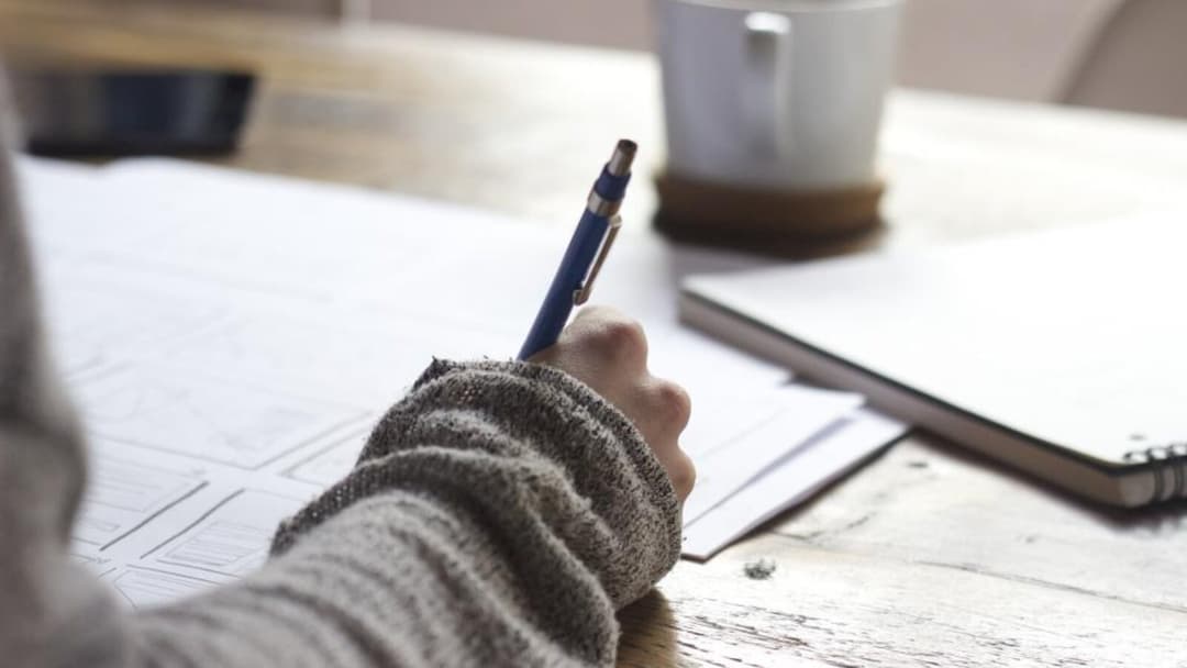 A hand in a knitted jumper writing with a blue pen on papers at a wooden desk in warm light, a white mug and a notebook beside it.