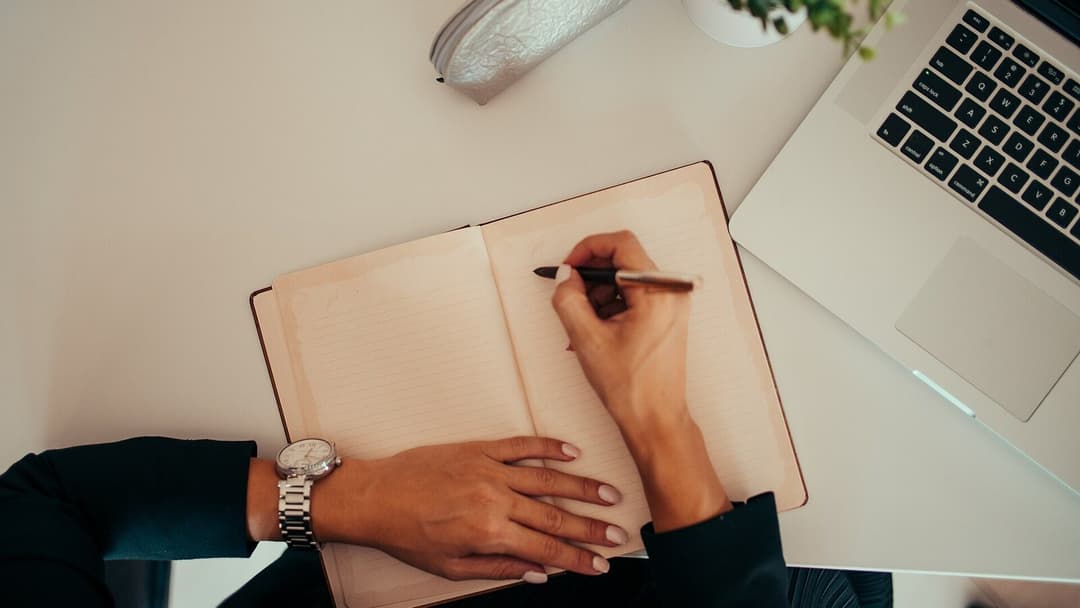 A top-down view of a person writing in an open lined notebook with a pen, a laptop and a small potted plant on a pale desk, a watch on the wrist.