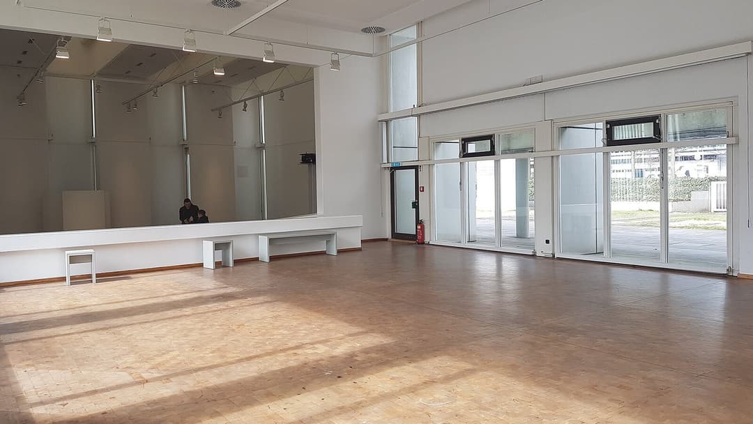 A bright, almost empty white gallery hall with a pale wooden parquet floor, track lighting overhead and tall glass doors opening to a paved area outside.