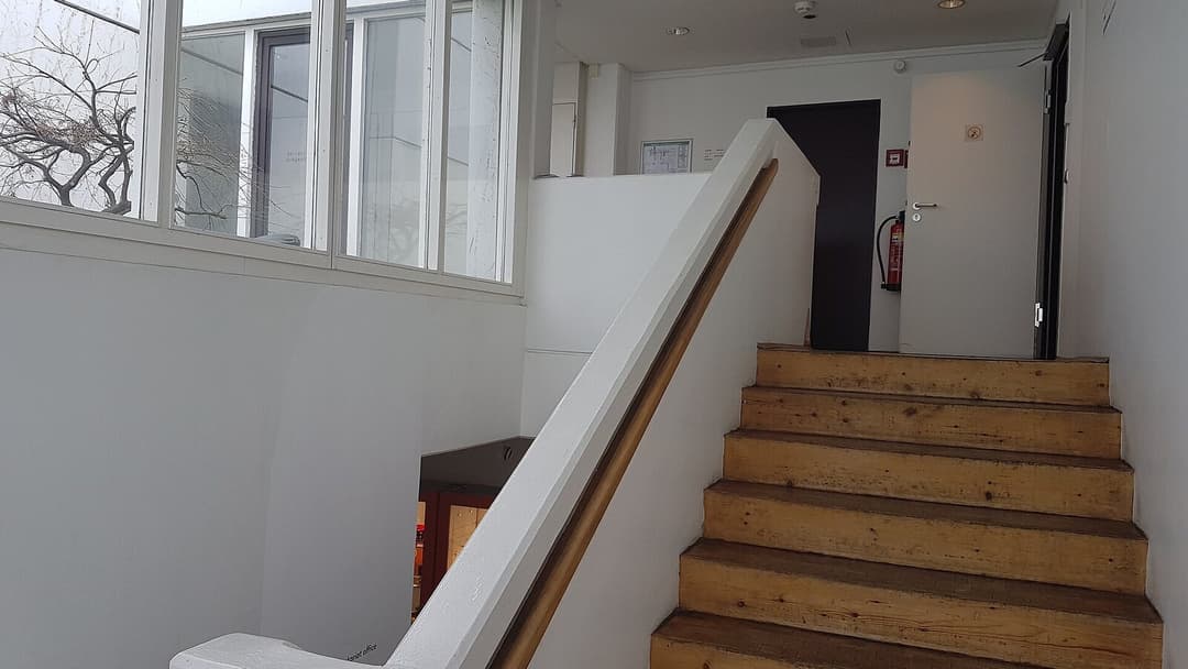 A plain white staircase with a slim wooden handrail and bare timber treads, lit by a bank of windows, with a dark doorway and a red fire extinguisher at the top.