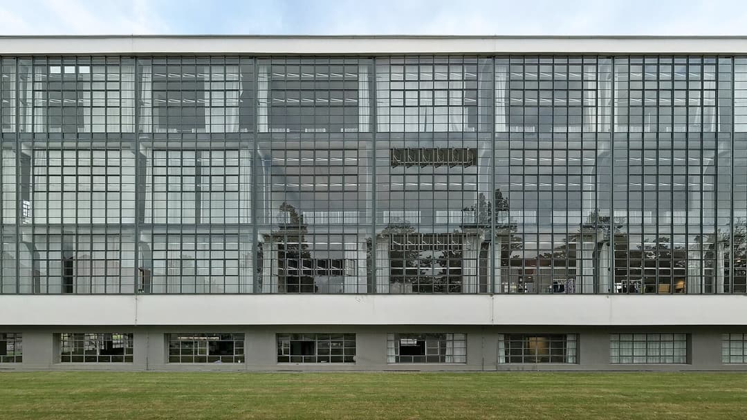 A large grid of slim steel-framed glass panels forming the curtain-wall front of a white building, with faint lettering visible behind the glass and a strip of grass along the base.