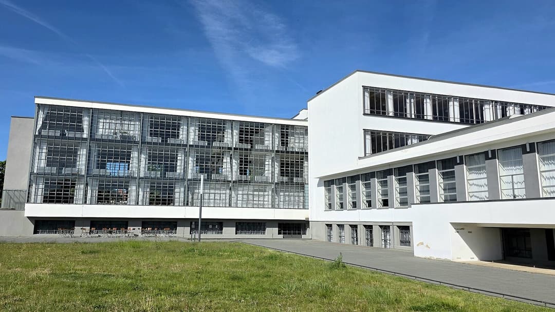 A long white modernist building with a glass-fronted workshop wing of ribbon windows on the left and a plainer white block on the right, set behind a lawn under a clear blue sky.