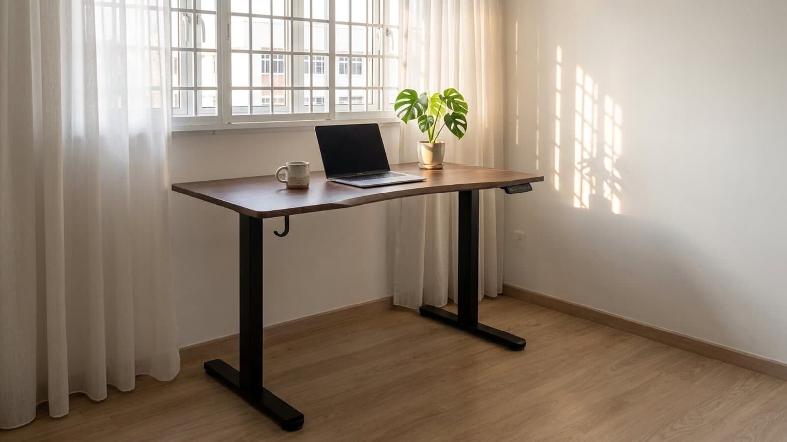 A walnut finish table on a black frame under a window in an HDB room, with a laptop, a mug and a monstera on top